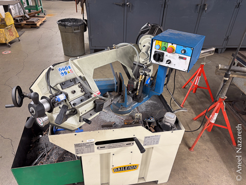 A bar being cut on a bandsaw set to a diagonal miter. Around the say you can see a bunch of test-cuts on tube trying to dial in the angle